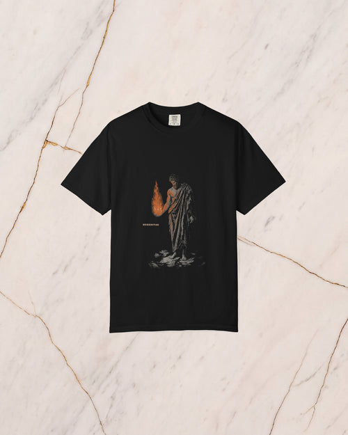 The PROMETHEUS shirt in black, laid on pale marble. Prometheus holds the stolen fire in his open hand, and its light falls on his forearm, his chest and the underside of his beard.