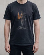 The PROMETHEUS shirt in black, worn, against a plain concrete wall. Prometheus holds the stolen fire in his open hand, and its light falls on his forearm and his chest.