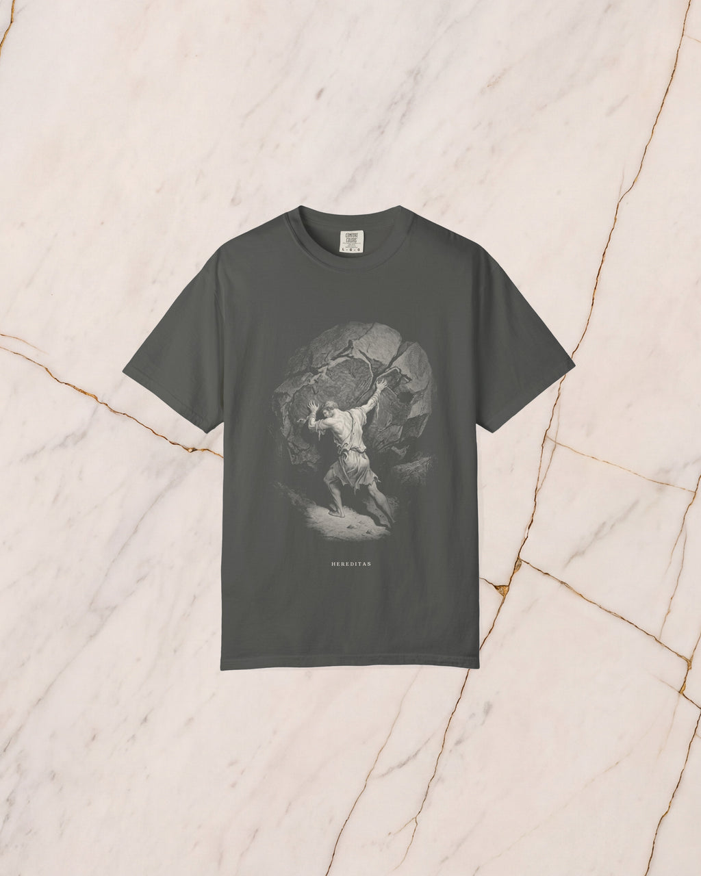 The SISYPHUS shirt in pepper, a dark warm charcoal, laid on pale marble. Sisyphus sets his shoulder to the boulder on the slope, drawn in bone line with no light in it but the day he works in.