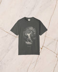 The SISYPHUS shirt in pepper, a dark warm charcoal, laid on pale marble. Sisyphus sets his shoulder to the boulder on the slope, drawn in bone line with no light in it but the day he works in.