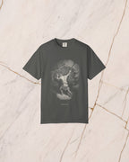 The SISYPHUS shirt in pepper, a dark warm charcoal, laid on pale marble. Sisyphus sets his shoulder to the boulder on the slope, drawn in bone line with no light in it but the day he works in.