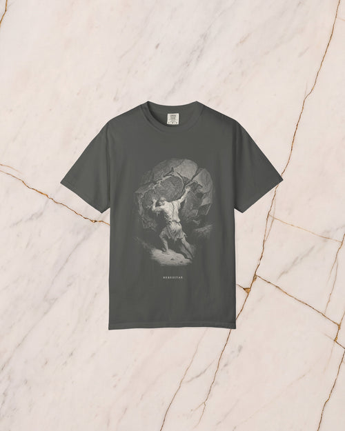 The SISYPHUS shirt in pepper, a dark warm charcoal, laid on pale marble. Sisyphus sets his shoulder to the boulder on the slope, drawn in bone line with no light in it but the day he works in.