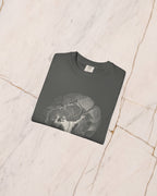 The SISYPHUS shirt in pepper folded on pale marble, showing the weight of the cotton and the detail of the artwork.