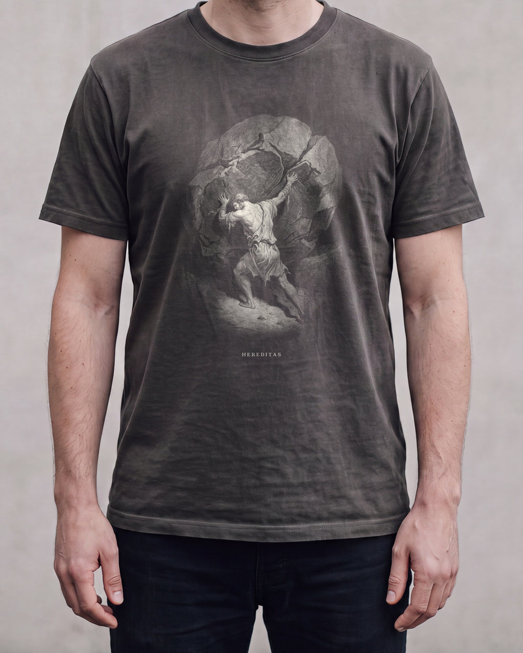 The SISYPHUS shirt in pepper, a dark warm charcoal, worn, against a plain concrete wall. Sisyphus sets his shoulder to the boulder, drawn in bone line.