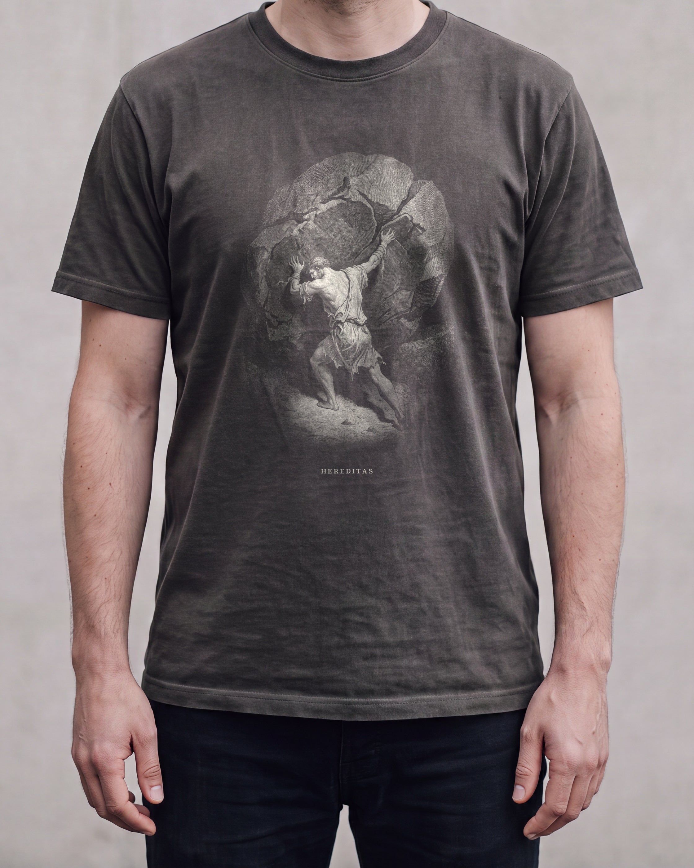 The SISYPHUS shirt in pepper, a dark warm charcoal, worn, against a plain concrete wall. Sisyphus sets his shoulder to the boulder, drawn in bone line.