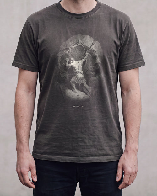 The SISYPHUS shirt in pepper, a dark warm charcoal, worn, against a plain concrete wall. Sisyphus sets his shoulder to the boulder, drawn in bone line.