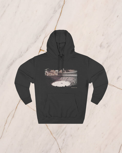 The VENATOR hoodie in charcoal heather, laid flat on pale marble. A hunter and a lion alone on the arena sand, the temples of the city rising beyond the wall, drawn in bone line and lit by a low sun.